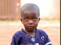 a young boy with blue eyes and a purple shirt is staring at the camera while standing in front of a building