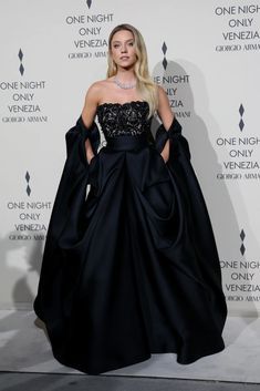 a woman standing in front of a white wall wearing a black dress with an off the shoulder design