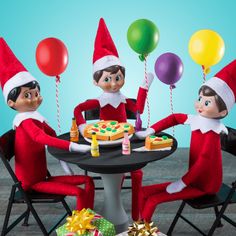 three elfs sitting at a table with pizza and balloons