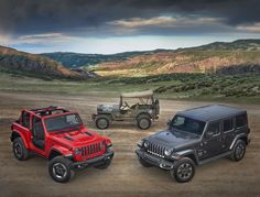 three jeeps are parked in the dirt near some hills and trees, one is red