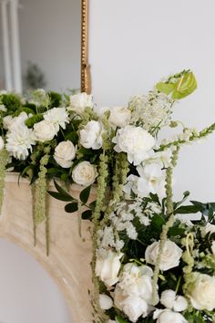 white flowers and greenery are arranged on the mantle in front of a gold mirror