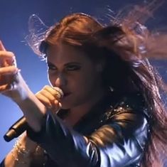 a woman holding a microphone in her right hand and wearing a leather jacket on stage