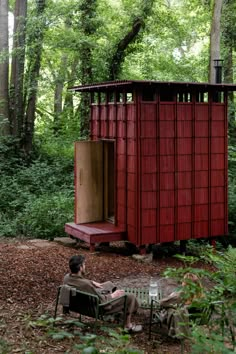 a man sitting on a chair in front of a red outhouse surrounded by trees