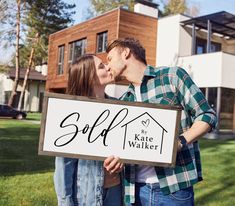 a man and woman kissing in front of a house while holding a sign that says sold by kate walker