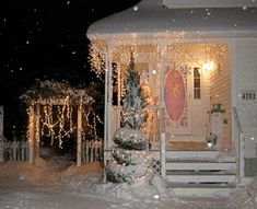 a house covered in snow with christmas lights on the front porch and trees around it