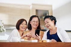 three women laughing while looking at a cell phone in the kitchen - stock photo - images