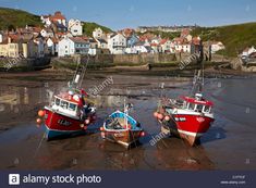 three fishing boats on the beach in front of some houses and cliffs, with low tide