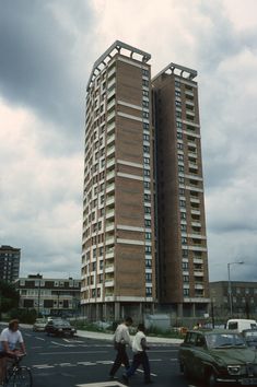 people crossing the street in front of two tall buildings on a cloudy day with cars and bicycles