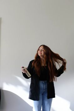 a woman with long red hair standing in front of a white wall and holding a cell phone