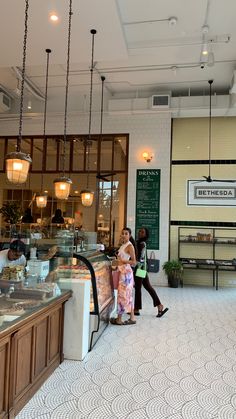 two people are walking in front of the counter at a bakery with lights hanging from the ceiling