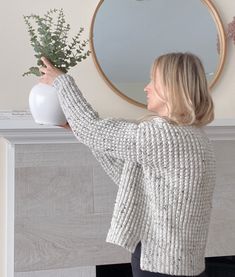 a woman holding a vase with flowers in it near a mirror on the wall above a fireplace