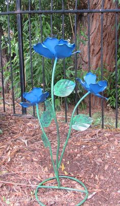 three blue flowers stand in front of a black iron fence and tree trunk with leaves on the ground