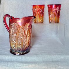 three red glass pitchers and two glasses on a white tableclothed cloth with gold trimmings