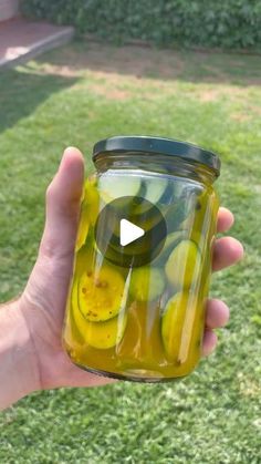 a hand holding a jar filled with pickles on top of a grass covered field