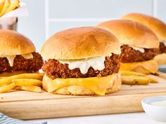 three fried chicken sandwiches with cheese and french fries on a cutting board next to some dipping sauce