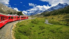 a red train traveling down tracks next to a lush green hillside covered in snow capped mountains