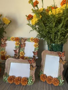 two frames with flowers and butterflies are on a table next to a potted plant