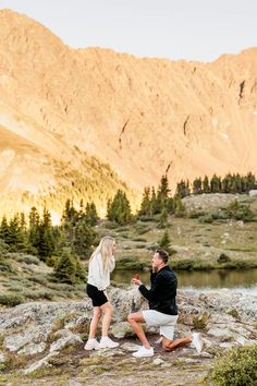 a man and woman sitting on top of a rock next to a lake with mountains in the background