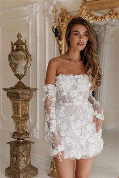 a woman wearing a white dress with flowers on the side and long sleeves, standing in front of a mirror