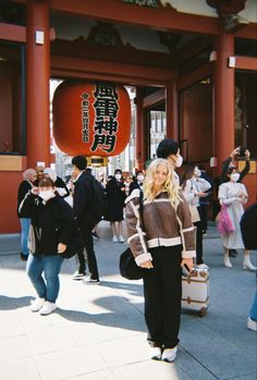 a woman standing in front of a red building with people walking around her and holding suitcases