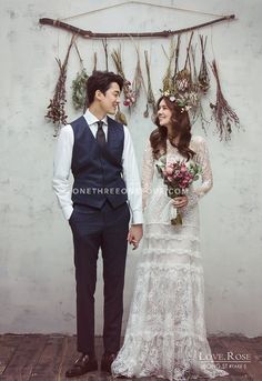 a man and woman standing next to each other in front of a wall with dried flowers