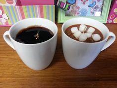 two white cups filled with hot chocolate and marshmallows on top of a wooden table