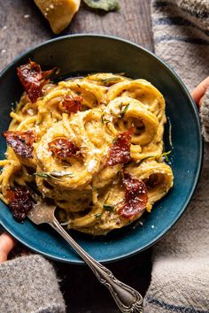 a bowl filled with pasta and bacon on top of a table next to a person's hand