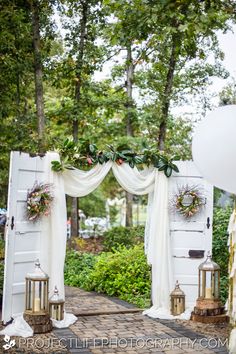 an outdoor wedding setup with white drapes and greenery on the door, surrounded by trees