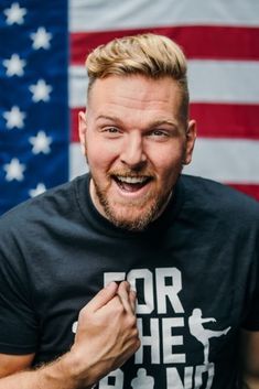 a man in front of an american flag making a funny face with his hand on his chest