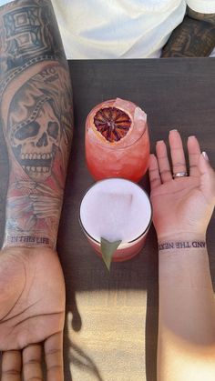 two people with tattoos on their arms sitting at a table and one has a drink in front of them