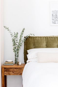 a bed with white sheets and pillows next to a wooden table with a potted plant on it