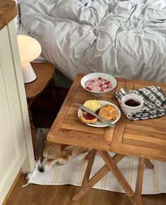 a dog eating food on a wooden table in front of a bed with white sheets