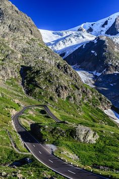 a winding road in the mountains with snow on the top and green grass around it