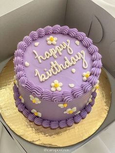 a birthday cake with purple frosting and white flowers on the top is in a box