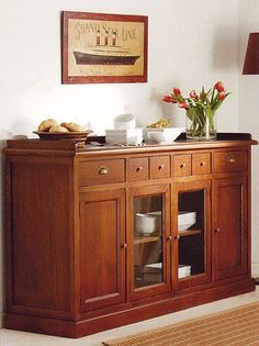 a wooden buffet table with glass doors and flowers on the top, in front of a white wall