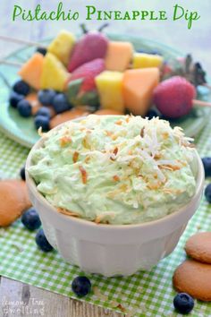 a white bowl filled with fruit salad next to cookies and other food on a green checkered tablecloth