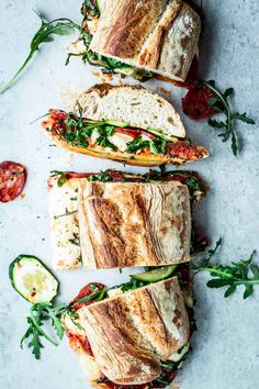 four sandwiches cut in half on top of a white surface with tomatoes, cucumbers and herbs