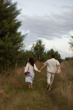 a man and woman holding hands walking down a path through tall grass with trees in the background