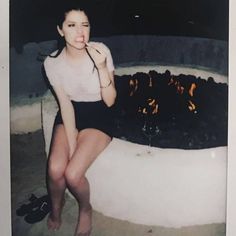 a woman sitting in front of a fire pit with her hand on her mouth and legs crossed