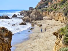 two people are walking along the beach by the water's edge, with cliffs in the background