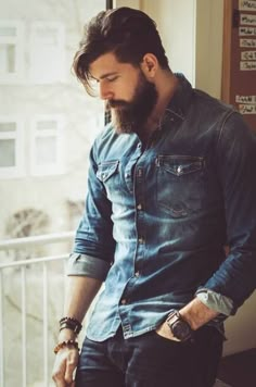 a man with a beard is looking down at his cell phone while standing in front of a window