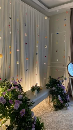 the room is decorated with white curtains and purple flowers on the floor, along with plants