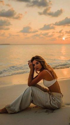 a beautiful woman sitting on top of a sandy beach next to the ocean at sunset