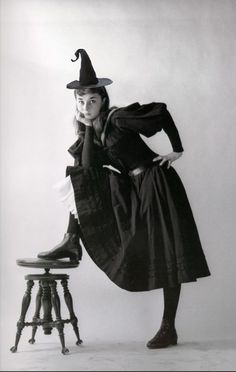 a black and white photo of a woman in a witch costume leaning on a stool