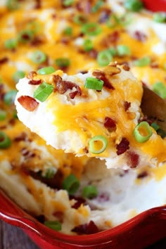 a red casserole dish filled with mashed potatoes, bacon and green onions