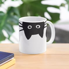 a white coffee mug with a black cat design on the front and side, sitting on a table next to a book