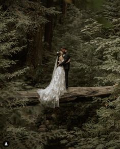 a bride and groom standing on a fallen log in the woods, hugging each other