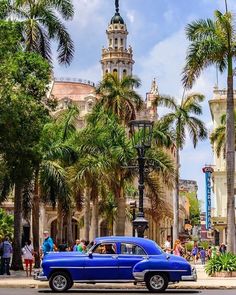 an old blue car parked in front of a tall building with a clock tower on top