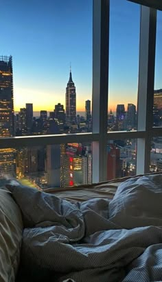 an unmade bed in front of a large window overlooking the city at night time