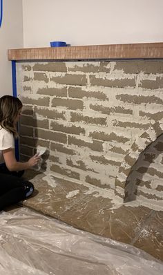 a woman sitting on the ground painting a brick wall with white paint and blue tape